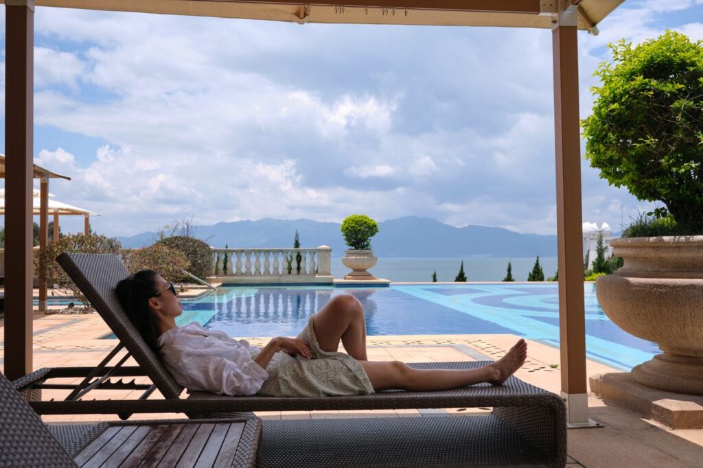 A woman in sunglasses relaxes on a sunlounger by a luxury poolside with a scenic view.